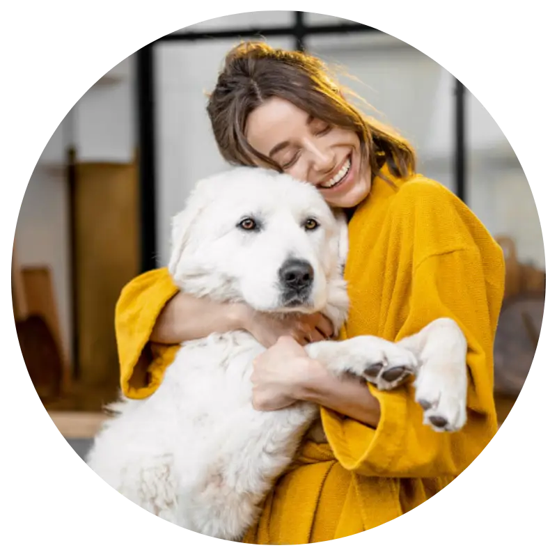 A person in a bright yellow robe holds a large, fluffy white dog with soulful eyes, set against a cozy indoor background.